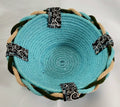 Turquoise woven bowl with black and white patterned straps on a white background